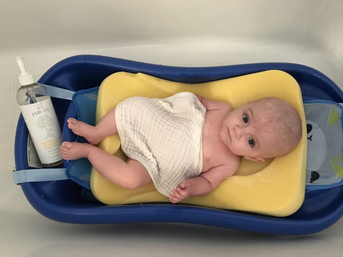 first years sure comfort deluxe baby tub with baby inside of it with a sponge sling and shampo caddy