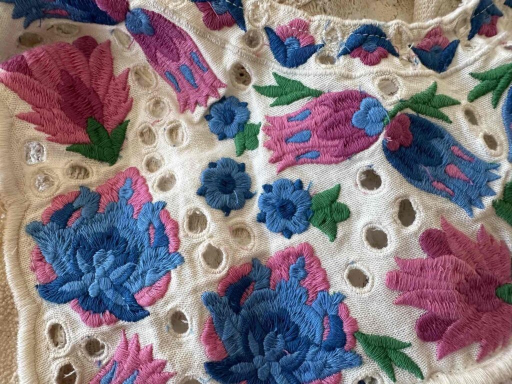 Close up of floral embroidery in pink, blue and green. Hand sewn on eyelet fabric